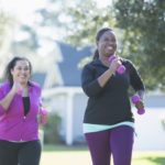 Two multi-ethnic women exercising outdoors, jogging on a sunny day in a residential neighborhood, handweights in their hands. The main focus is on the African American woman, laughing as her Hispanic friend tries to catch up with her.