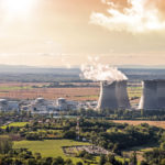Horizontal composition photography of french nuclear power station with four steaming cooling towers in countryside plain with smoke cloud (water condensation). Image taken from high angle view, aerial view, in Bugey, in Ain on the border of Isere department, Rhone-Alpes region in France (Europe). The nuclear power station is located in the middle of a plain landscape in France, near Lyon city. This picture was taken during a bright orange sunset in autumn season with green and brown meadow and field.