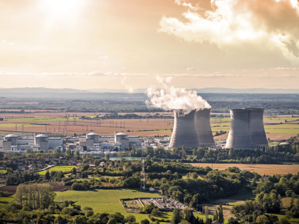 Horizontal composition photography of french nuclear power station with four steaming cooling towers in countryside plain with smoke cloud (water condensation). Image taken from high angle view, aerial view, in Bugey, in Ain on the border of Isere department, Rhone-Alpes region in France (Europe). The nuclear power station is located in the middle of a plain landscape in France, near Lyon city. This picture was taken during a bright orange sunset in autumn season with green and brown meadow and field.