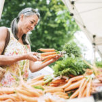 An older woman in her fifties shopping in a local farmers market with fresh, organic vegetables. She smiles as she chooses carrots. Horizontal image with copy space.