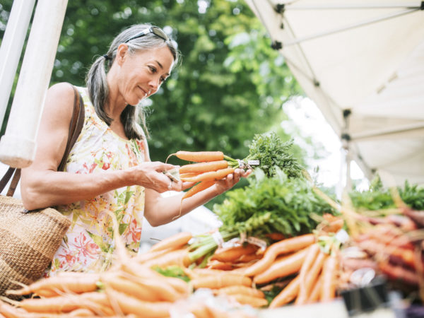An older woman in her fifties shopping in a local farmers market with fresh, organic vegetables. She smiles as she chooses carrots. Horizontal image with copy space.