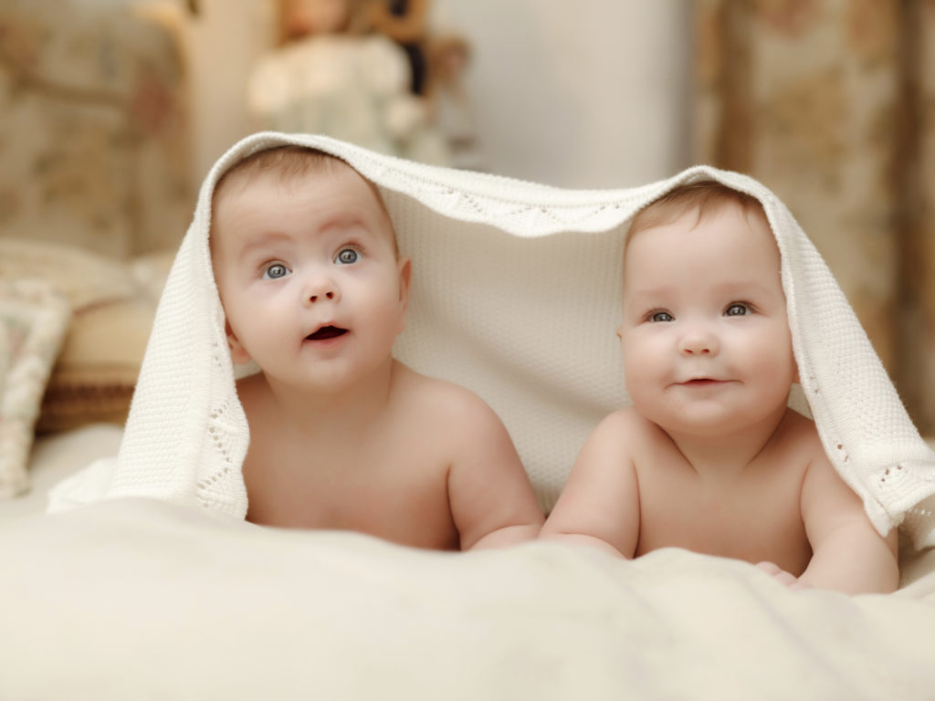 Two twin babies, seven-month smiling girls covered with a white towel in bed on white sheets