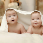 Two twin babies, seven-month  smiling girls covered with a white towel in bed on white sheets