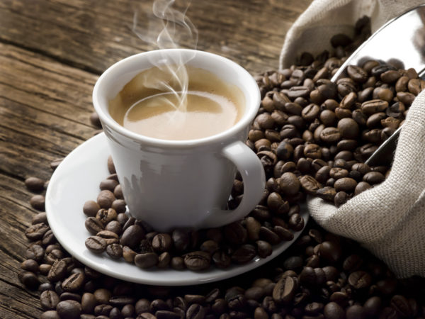 Steaming hot coffee in a small white coffee cup sits on a round saucer and is surrounded by coffee beans.  A sack of coffee beans with a shiny metal coffee scooper sticking out of it is visible along the right side of the image.  Beans are spilling from the sack onto the saucer and the brown wooden table beneath it.