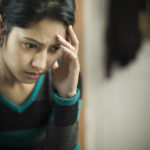 Indoor low key image of a serene, stressed young woman feeling headache and thinking by touching her head and looking down with blank expression. She is wearing a sweater. One person, waist up, horizontal composition with selective focus and copy space.