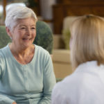 Front view of smiling senior patient sitting in armchair talking with home healthcare nurse or female doctor, rear view.