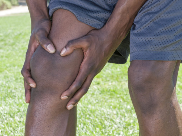 Closeup of knee and leg of lean African American male athlete clutching injured knee with fingers around the patella on green lawn outdoors