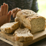 A woman rejecting bread, gluten-free concept. A whole-grain loaf of bread on a rustic wooden table, and a woman rejecting it with a hand gesture.