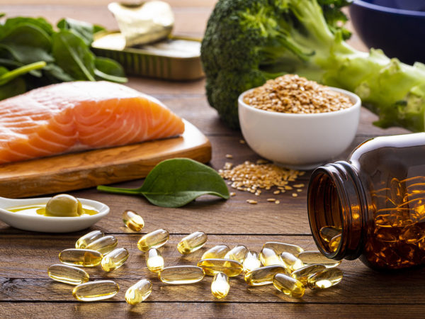 Front view of many fish oil capsules spilling out from the bottle surrounded by an assortment of food rich in omega-3 such as salmon, flax seeds, broccoli, sardines, spinach, olives and olive oil.