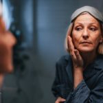 Portrait of a beautiful older woman in her pajamas and with a sleeping mask on her forehead, looking at her reflection in the mirror and checking her face and wrinkles.