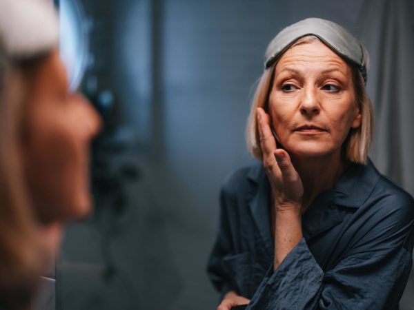 Portrait of a beautiful older woman in her pajamas and with a sleeping mask on her forehead, looking at her reflection in the mirror and checking her face and wrinkles.